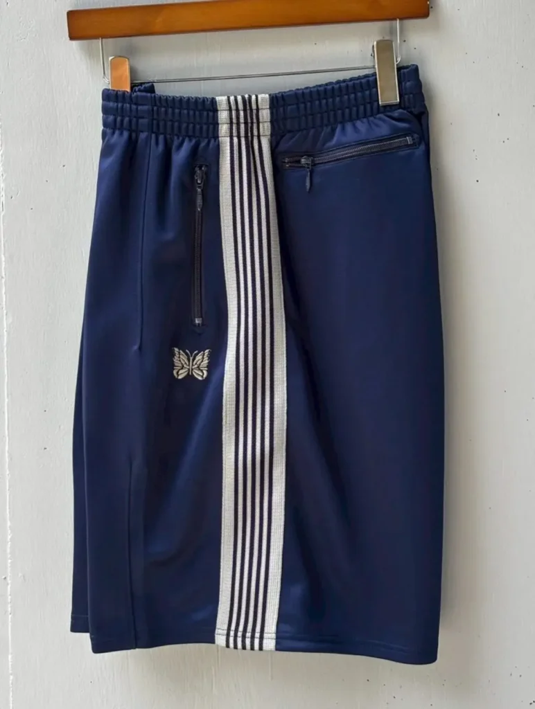 Navy blue Needles track shorts hanging on a wooden hanger with a white striped center panel, zip pockets, elastic waistband, and embroidered butterfly logo on the left leg