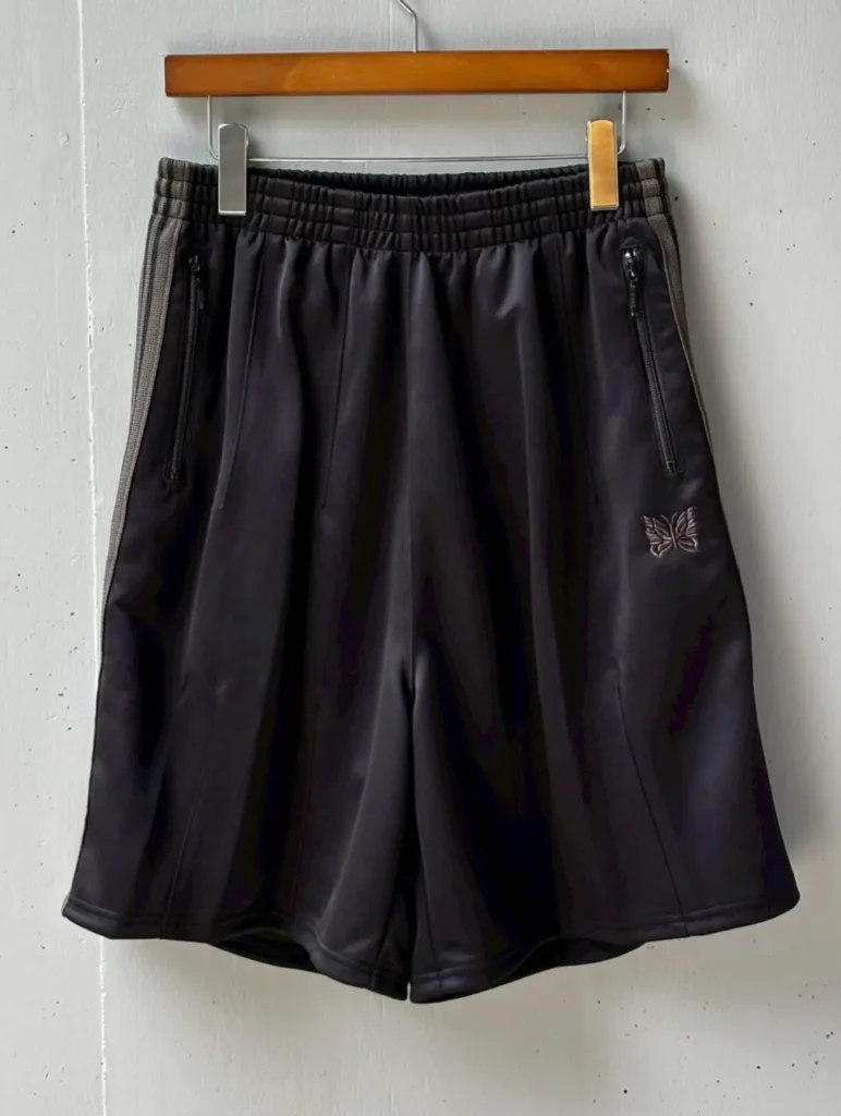 Black Needles track shorts shown from the front on a hanger. Olive side stripes, dual zip pockets, elastic waistband, and the embroidered butterfly logo create a clean athletic look