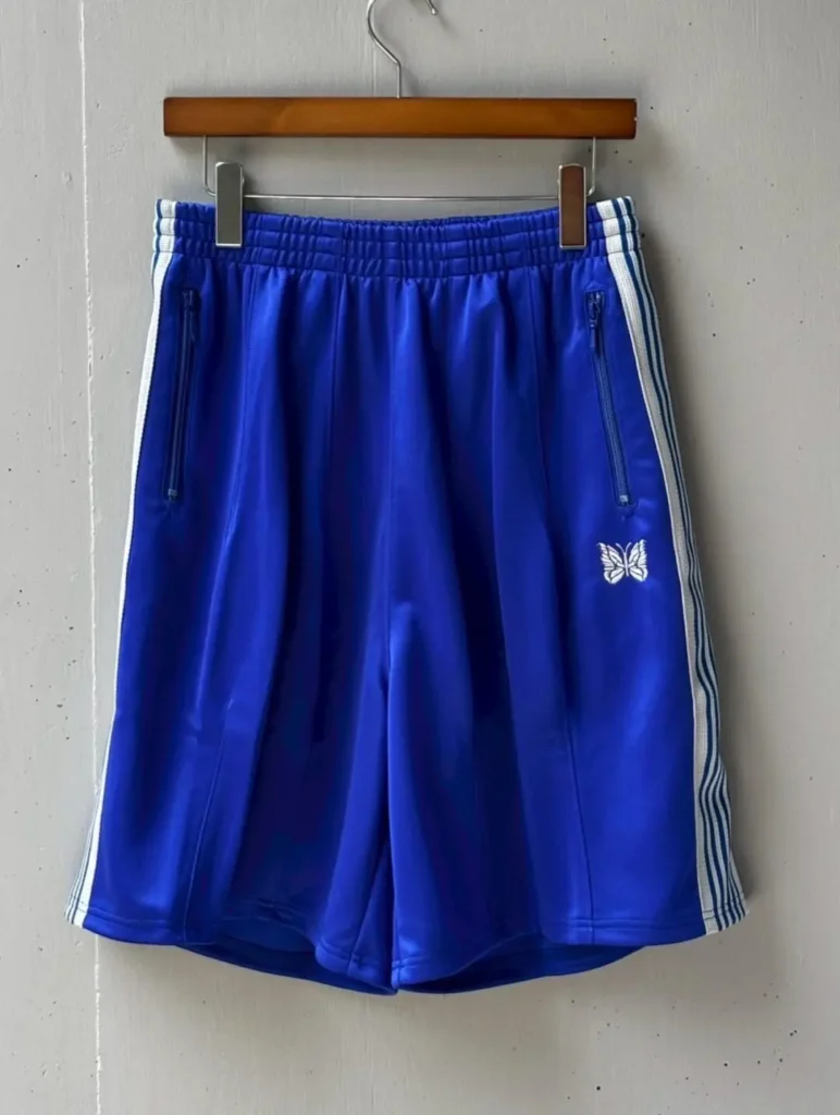 Front view of royal blue Needles track shorts featuring white side stripes, zip pockets, elastic waistband, and a white embroidered butterfly logo near the lower right leg
