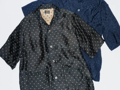 Two relaxed-fit short-sleeve camp-collar shirts from Needles displayed on a light gray background. The front shirt features a black jacquard fabric with small beige polka dots, oversized buttons, and large patch pockets. Layered behind it is a navy blue version with a tonal textured pattern, showcasing the brand’s signature loose silhouette and luxurious drape