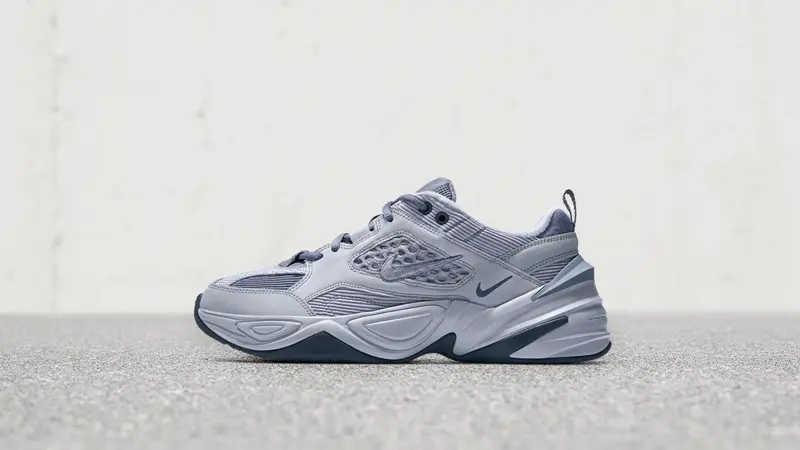 Side-profile product shot of a Nike M2K Tekno-style shoe in cool grey tones. The shoe features layered mesh and synthetic overlays, perforated side panel detailing, a small tonal Swoosh, and a sculpted chunky midsole with darker grey accents. Photographed against a minimalist concrete backdrop