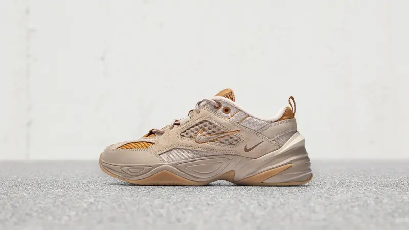 Side-profile view of a Nike lifestyle runner dressed in soft beige and tan hues. Textured mesh underlays, suede-like overlays, and metallic gold accents combine with a sculpted midsole and subtle Swoosh branding. Captured in a clean studio setting with a neutral background