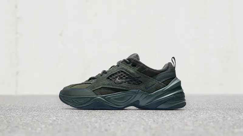 Side-profile product image of a Nike shoe in deep olive and black tones. The design combines rugged mesh, layered overlays, perforated side panels, and tonal branding with a bold geometric midsole. Set against a minimalist grey backdrop