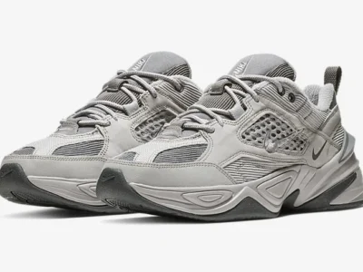 Side-profile product image of a Nike shoe in deep olive and black tones. The design combines rugged mesh, layered overlays, perforated side panels, and tonal branding with a bold geometric midsole. Set against a minimalist grey backdrop