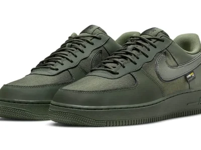 Olive green Nike Air Force 1 Low shoe shown in a three-quarter front view. The pair features durable Cordura fabric panels, smooth synthetic overlays, tonal laces, a dark olive Swoosh, matching rubber sole, and a small Cordura fabric tag near the heel
