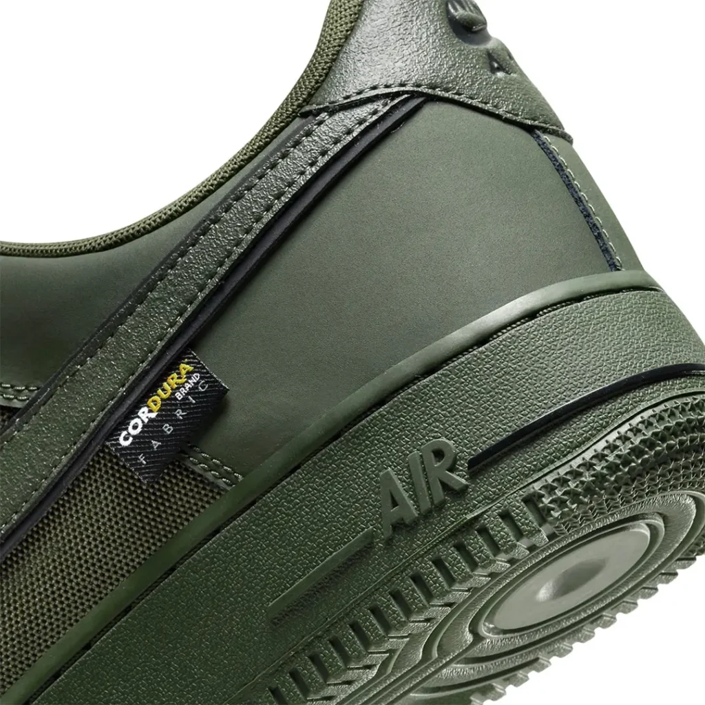 Close-up of the lateral heel showing the Cordura fabric tag stitched beneath the olive Nike Swoosh. The textured midsole features embossed "AIR" branding, highlighting the shoe's durable construction and monochrome finish