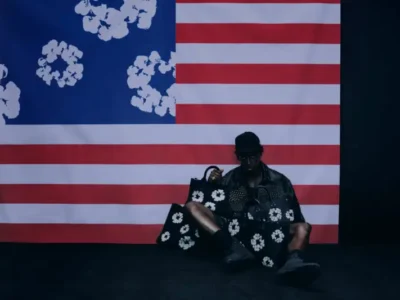 Model seated on the floor in front of a reimagined American flag backdrop, surrounded by oversized black leather bags featuring Denim Tears’ signature white cotton wreath motif. The moody lighting emphasizes the collection’s bold graphic contrast and cultural symbol