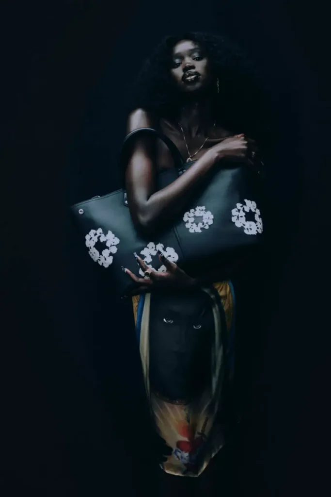 Studio portrait of a model holding a large black leather tote bag featuring white cotton wreath graphics. The dark backdrop and low-key lighting draw attention to the accessory’s texture, craftsmanship, and signature brand