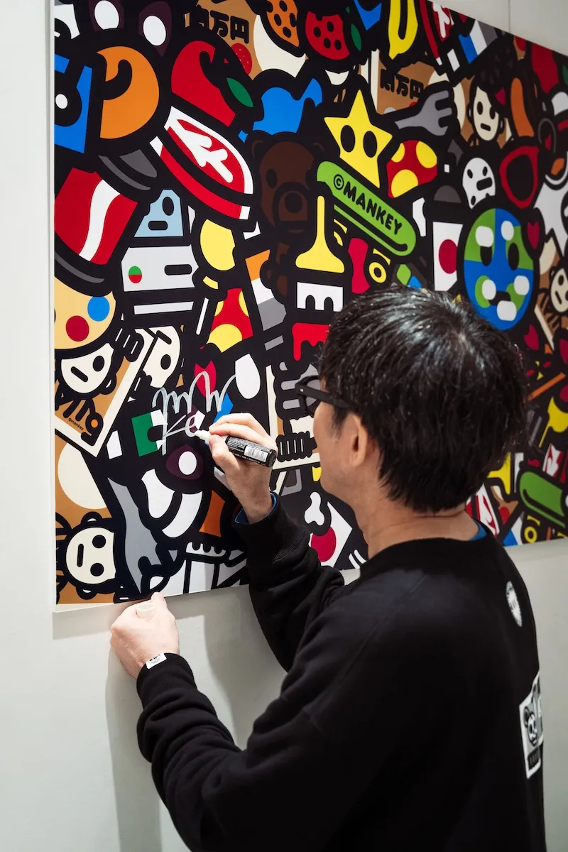 A person signs a vibrant contemporary artwork mounted on a gallery wall, using a marker to add their signature to the lower section of the piece. The canvas is filled with dense cartoon-inspired graphics, playful characters, gaming references, symbols, and bold geometric shapes rendered in black, red, blue, yellow, green, and white. Captured from behind, the scene highlights the artist’s interaction with the work, emphasizing its energetic pop-art aesthetic and intricate visual storytell