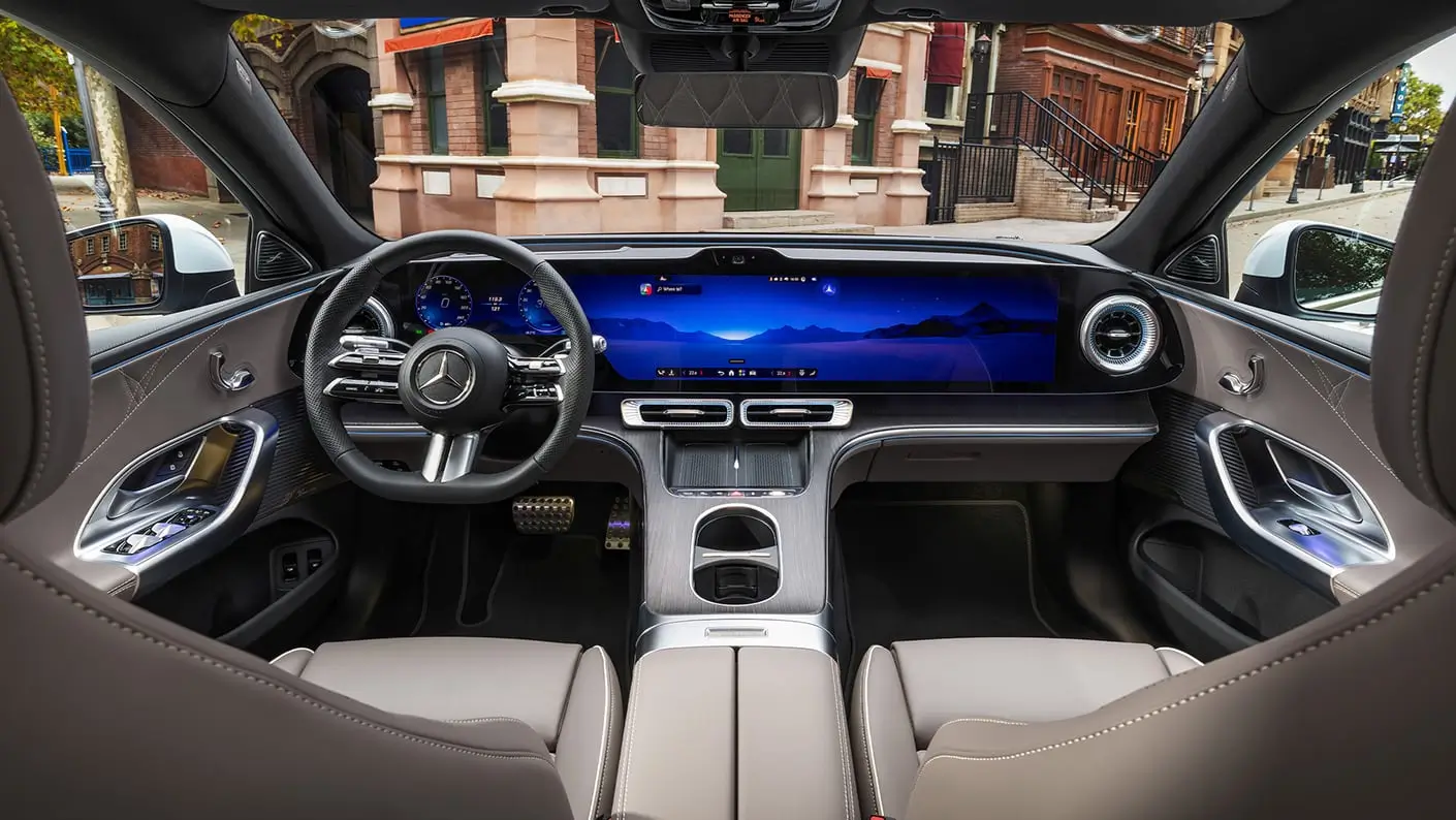 Wide-angle view of the Mercedes-Benz GLC electric SUV interior from the rear seats, showcasing a panoramic dashboard dominated by a seamless digital display spanning the instrument cluster and infotainment system. The cabin features taupe leather upholstery with contrast stitching, metallic trim, turbine-style air vents, ambient blue lighting, and a minimalist center console. Through the windshield, a row of historic urban brownstone buildings frames the upscale, technology-focused cockpit