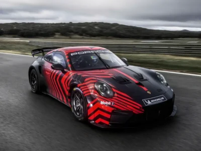 Porsche 911 GT4 R prototype speeding around a racetrack, finished in matte black with striking red camouflage graphics, large rear wing, Michelin branding, and aggressive aerodynamic bodywork under overcast skies