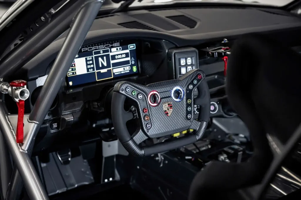 Race-prepared cockpit of the 2027 Porsche 911 GT4 R featuring a carbon-fiber steering wheel with integrated controls, digital instrument display, roll cage, racing seat, and simplified motorsport dashboard