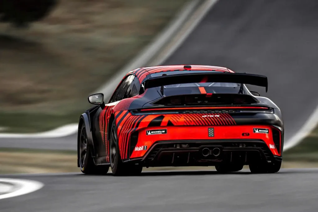 Rear three-quarter action shot of the 2027 Porsche 911 GT4 R cornering on a circuit, highlighting its full-width LED taillight, oversized rear wing, rear diffuser, dual exhaust outlets, and red camouflage livery