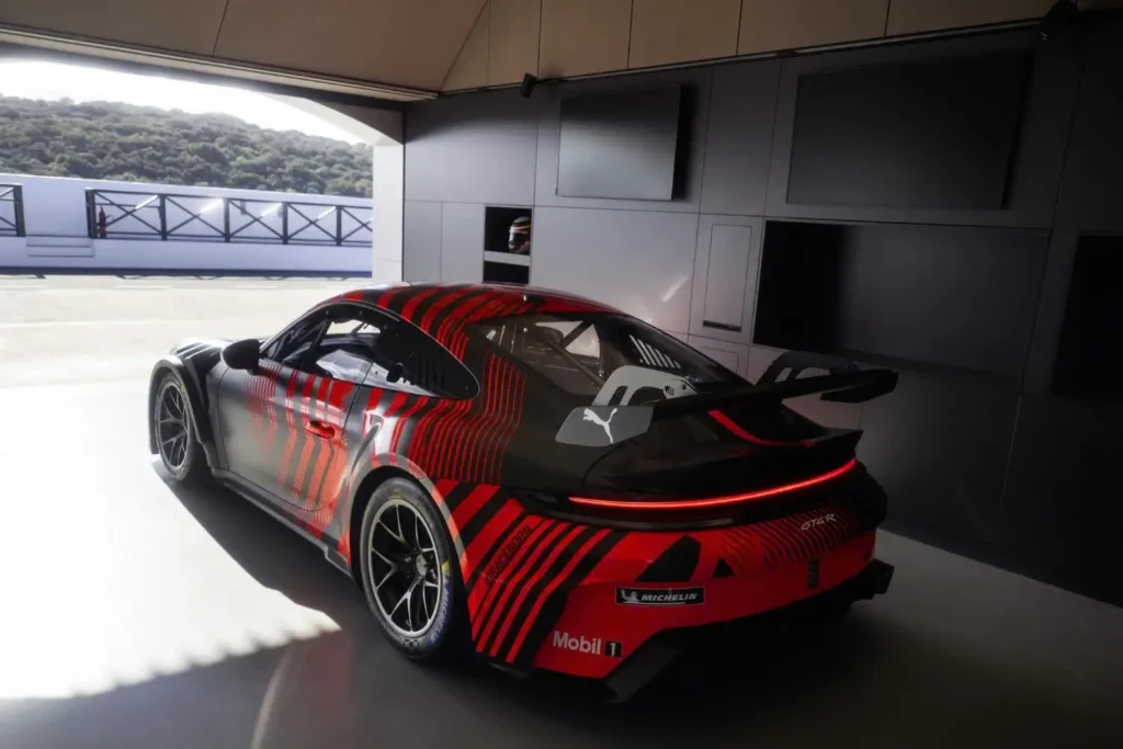Rear three-quarter view of the 2027 Porsche 911 GT4 R parked inside a pit garage, showcasing its aerodynamic rear wing, sculpted bodywork, lightweight wheels, and bold red-and-black camouflage wrap