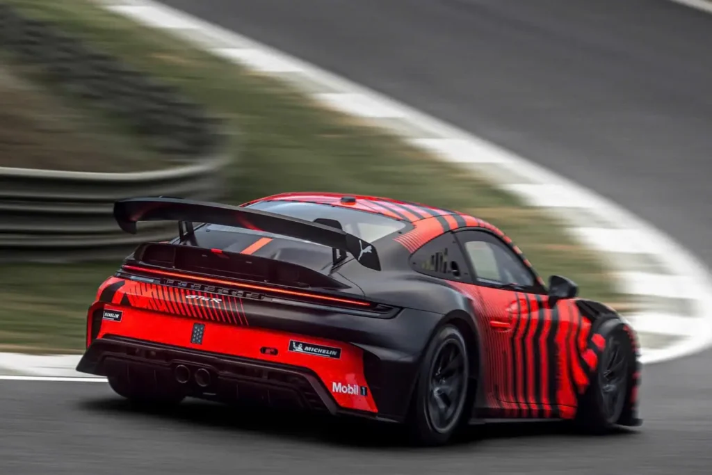 Rear three-quarter view of the 2027 Porsche 911 GT4 R prototype speeding through a racetrack corner, featuring a black-and-red camouflage livery, oversized rear wing, full-width LED taillight, aggressive rear diffuser, dual center exhausts, and wide racing stance in motion