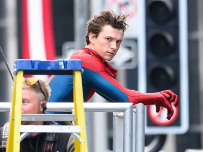 A behind-the-scenes set photo showing an actor in a red-and-blue Spider-Man costume leaning against a metal platform during filming. A yellow ladder partially obscures the foreground, while traffic lights, street signage, and production equipment create an urban backdrop. The candid moment captures the superhero character between takes, offering a glimpse into the filmmaking process on a city location set