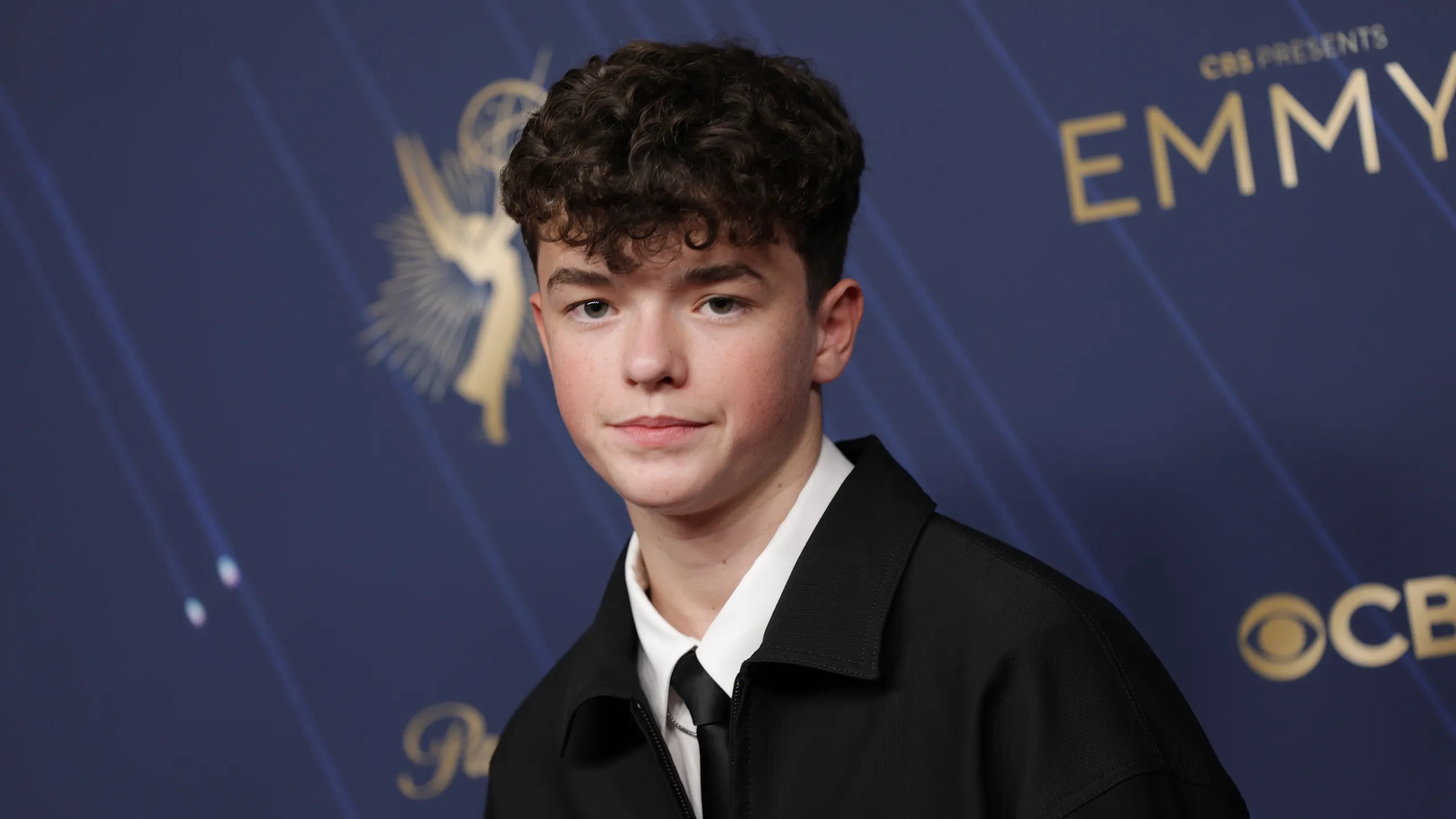 A portrait of a young attendee posing on the red carpet at a major television awards event. Wearing a black jacket, white shirt, and slim black tie, the individual stands before a navy-blue step-and-repeat backdrop featuring Emmy Awards branding and sponsor logos. Soft event lighting and a shallow depth of field create a polished, professional awards-show atmosphere