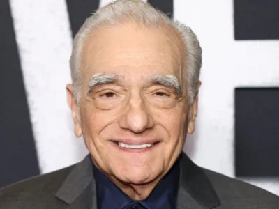Close-up portrait of an elderly man with silver hair and thick white eyebrows smiling at a red carpet event. He wears a dark charcoal suit with a navy shirt and tie, standing in front of a black-and-white promotional backdrop