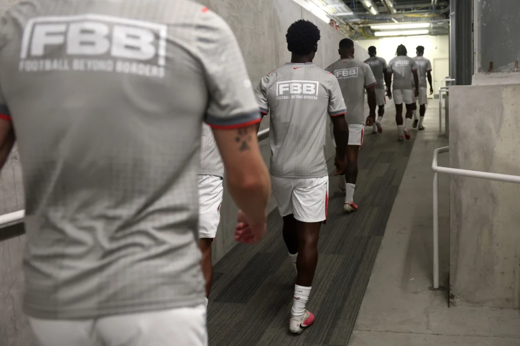 A group of young football participants wearing grey Football Beyond Borders (FBB) jerseys walk through a stadium tunnel toward the pitch. The industrial concrete corridor is illuminated by overhead lights, creating a sense of anticipation and teamwork as the players head toward an event or match experience