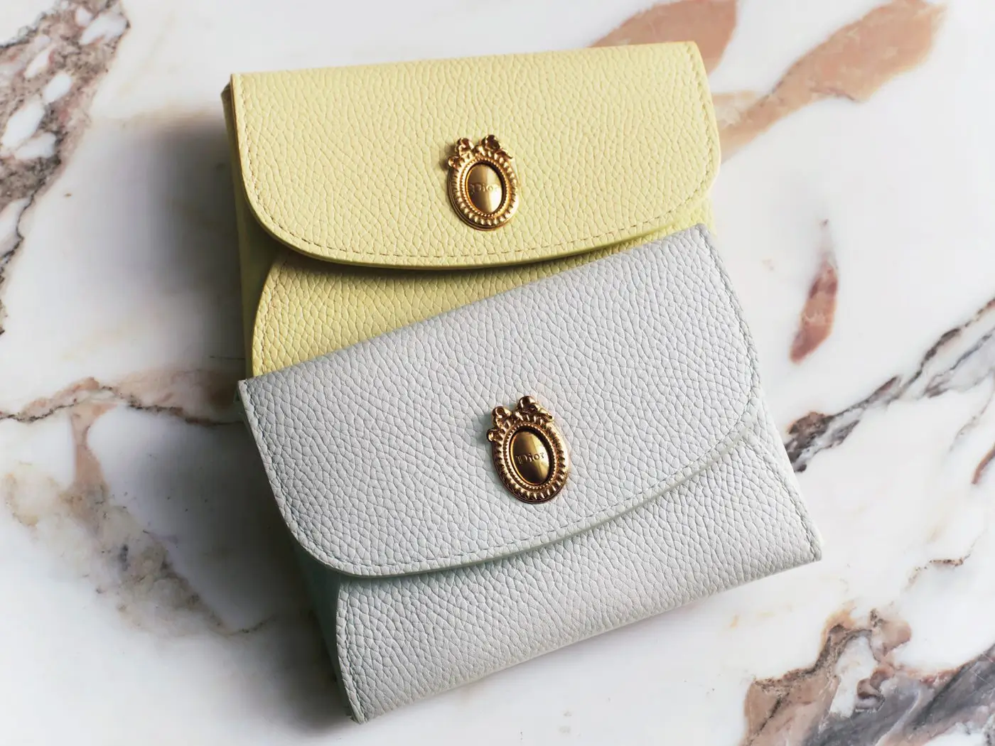 Two Dior Médaillon leather card holders displayed on a polished marble surface, featuring grained calfskin construction in soft pastel yellow and pale mint tones. Each piece is accented with a gold-tone Médaillon clasp inspired by Dior's historic rococo motif, blending elegant craftsmanship with refined day functionality