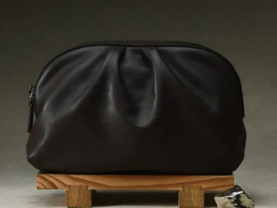 Dark brown leather pouch with sculptural gathered detailing and zip closure displayed on a small wooden stand in a minimalist studio set