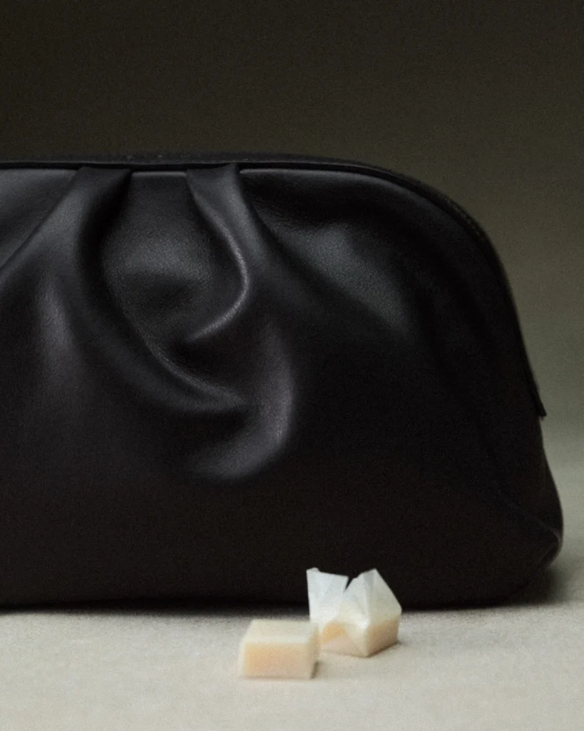 Cropped view of a dark leather pouch with sculpted folds and smooth matte finish beside small geometric stone accent