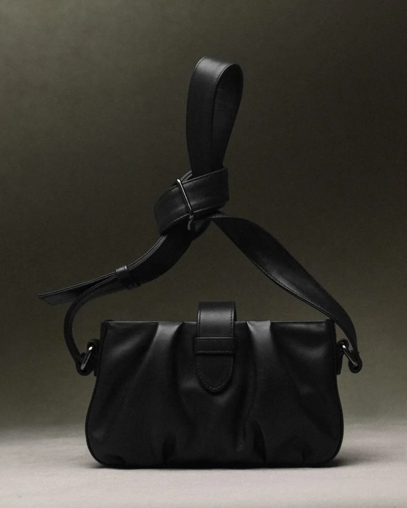 Black leather shoulder bag with wide strap and sculptural draped front showcased against a muted studio backdrop