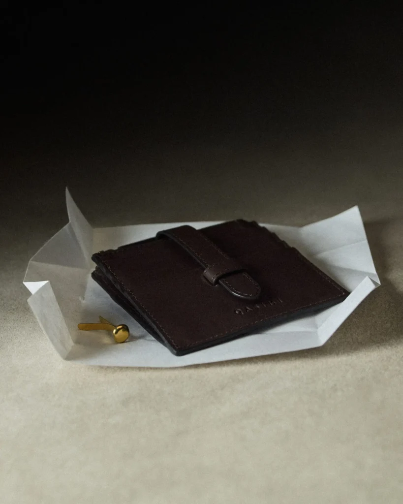 Compact dark brown leather card holder with strap closure displayed on folded paper, showcasing clean lines and understated craft