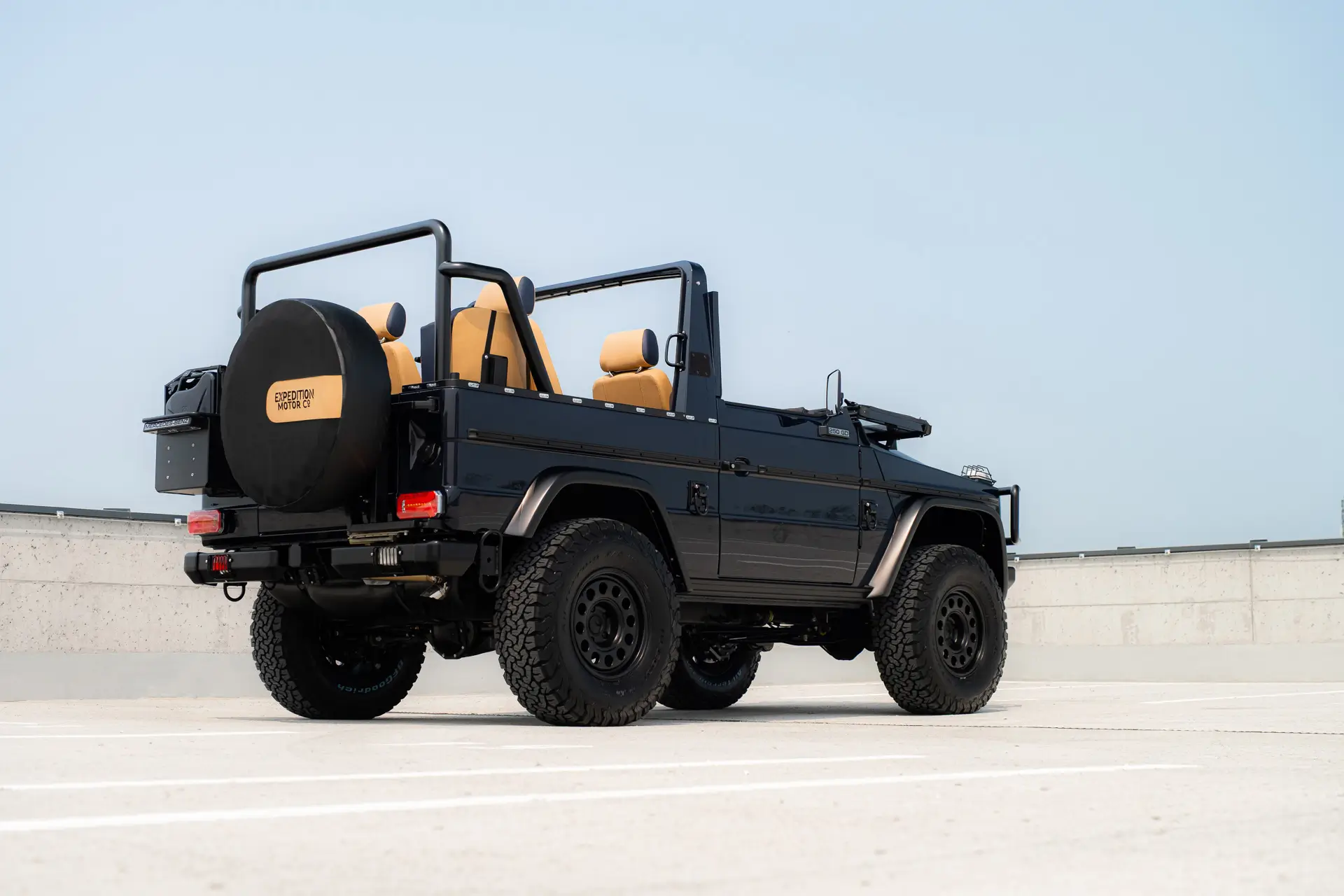 Rear three-quarter view of a black Expedition Motor Company vintage cabrio G-Wagen parked on a rooftop deck, showcasing its open-air body, tan leather seats, rugged off-road tires, and classic short-wheelbase proportion