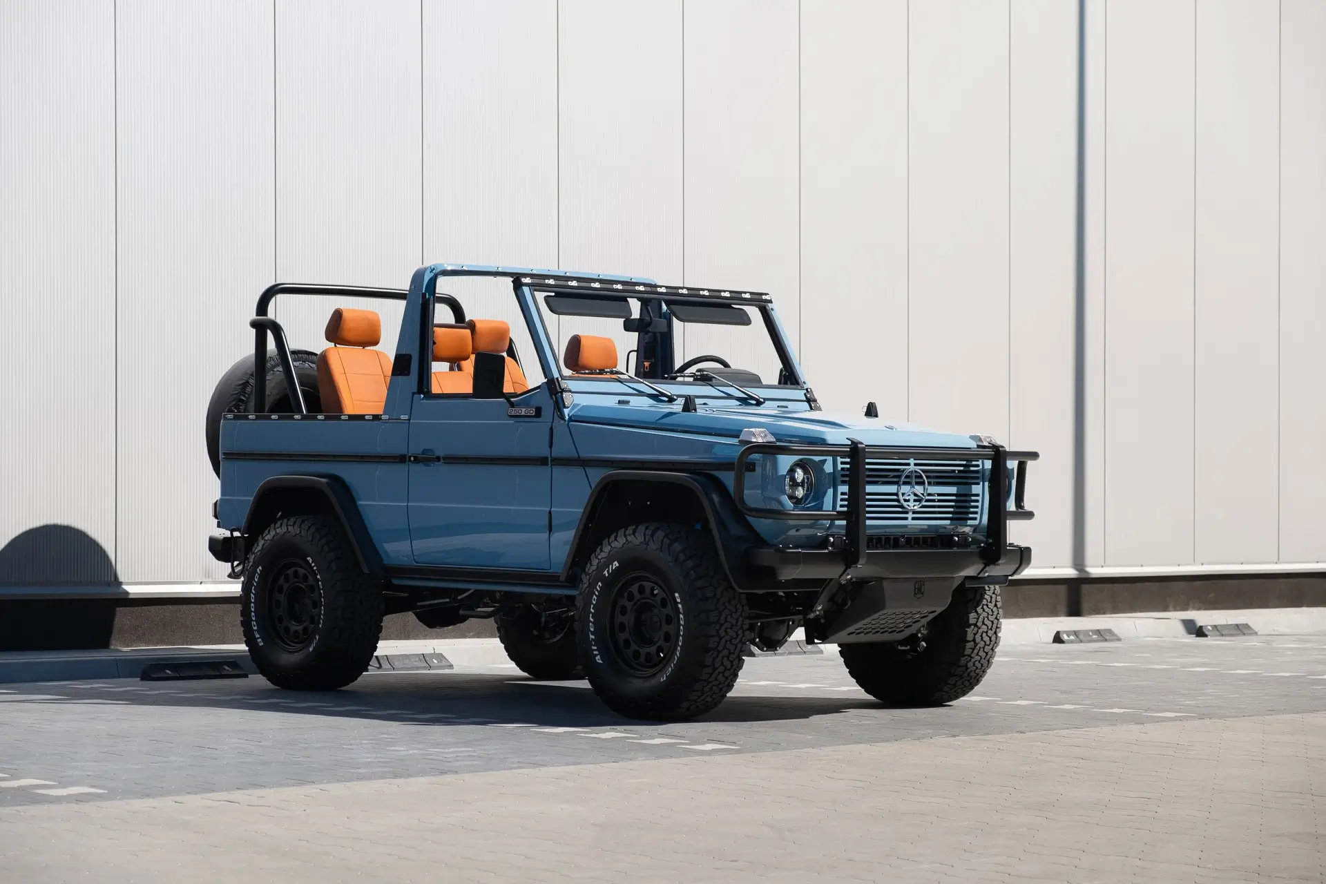 Front three-quarter view of a light blue Mercedes-Benz 250GD Wolf cabriolet restored by Expedition Motor Company, featuring an open-top configuration, military-inspired brush guard, and orange leather interior accent