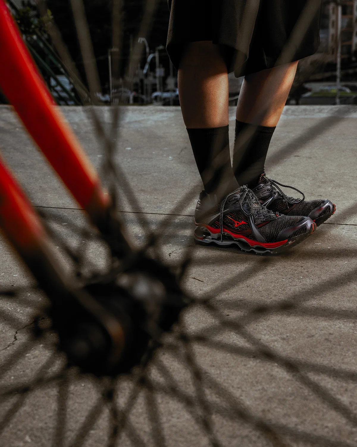 Lifestyle shot of the Mizuno Wave Prophecy sneaker in a black and red colorway, worn with black shorts and crew socks on an urban pavement surface. A blurred bicycle wheel and red bike frame dominate the foreground, creating depth while spotlighting the shoe’s mesh upper, metallic overlays, and signature Wave sole cushion