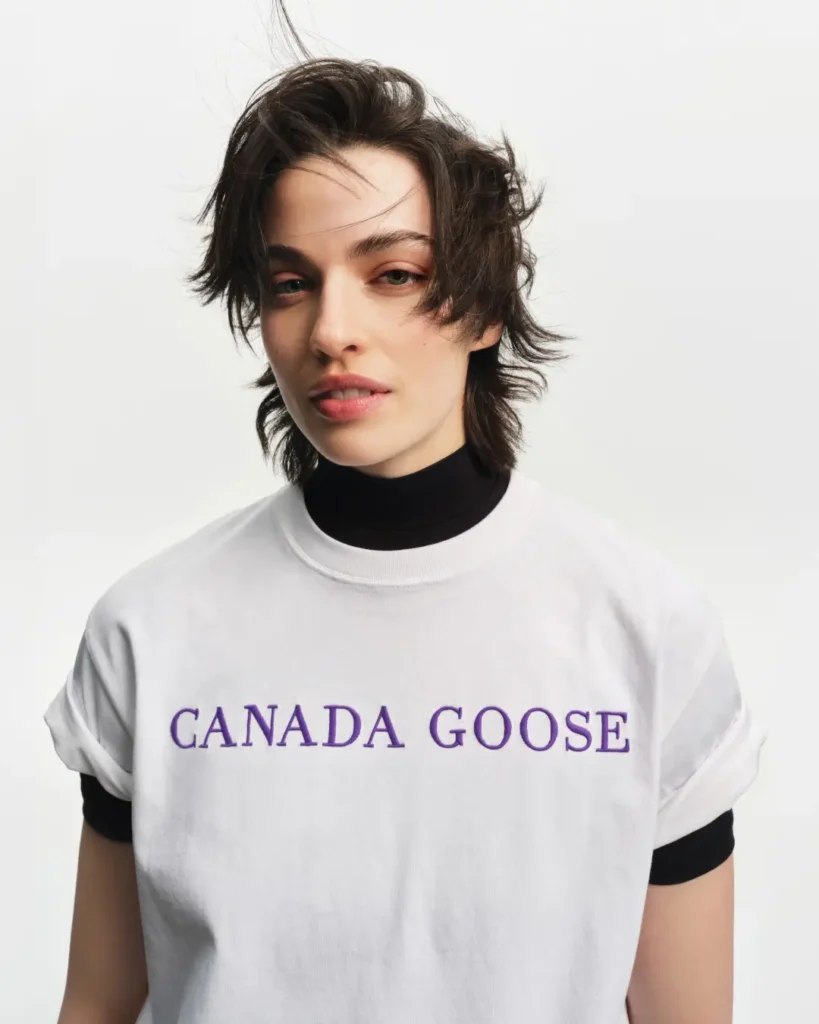 Portrait of a model wearing a white Canada Goose logo T-shirt layered over a black mock-neck top from the Summer 2026 collection