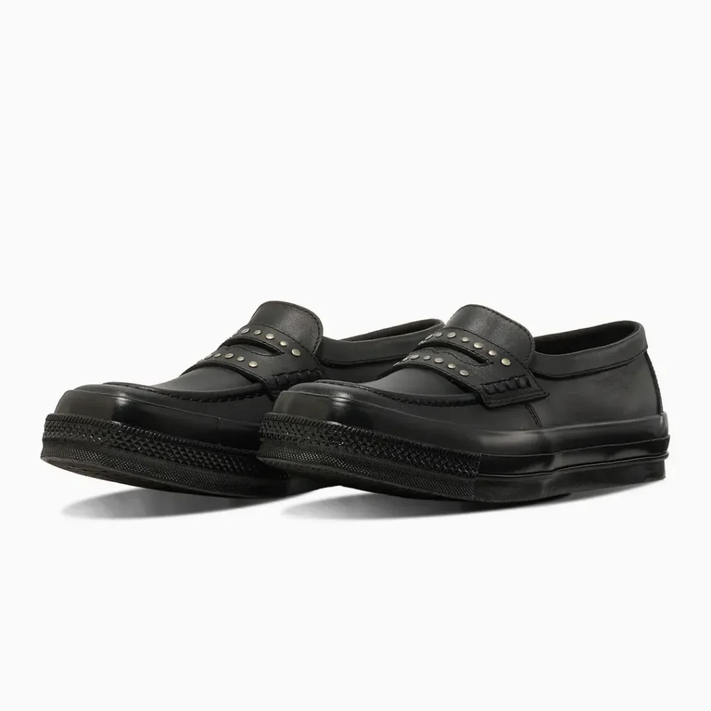 Pair of black leather Converse penny loafers shown from a front three-quarter angle, highlighting the studded strap, moc-toe stitching, and textured black shoe