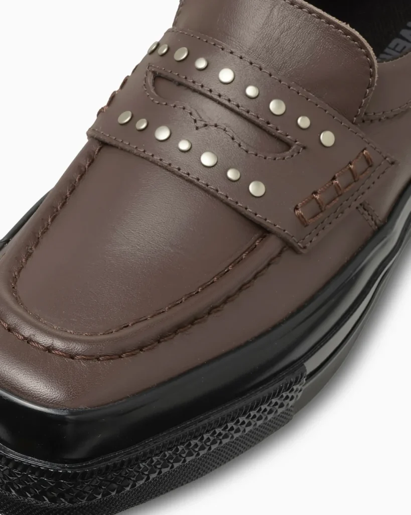 Close-up of the brown leather upper emphasizing the silver metal studs, penny strap, moc-toe stitching, and smooth leather texture
