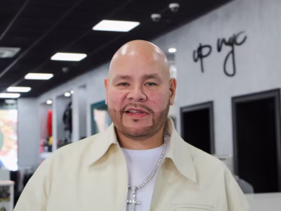 Portrait of a man wearing a cream-colored jacket and diamond cross necklace inside a modern retail space, standing in front of a minimalist interior with clothing displays and overhead light