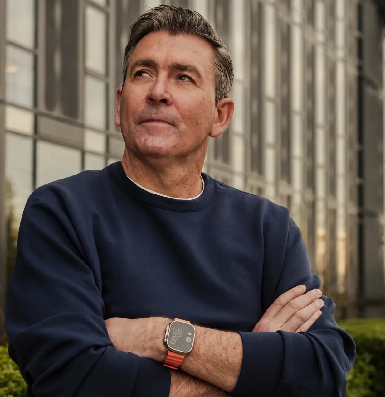 Portrait of Phil McCartney wearing a navy crewneck sweatshirt and orange-banded smartwatch, standing with arms crossed outside a modern office building, looking thoughtfully into the distance in a professional leadership profile photograph