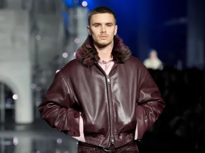 A fashion model walks down a runway wearing a deep burgundy leather bomber jacket with a plush fur collar, paired with matching dark-toned garments. The oversized silhouette and rich textures are highlighted by dramatic runway lighting, emphasizing a luxurious and contemporary menswear aesthetic