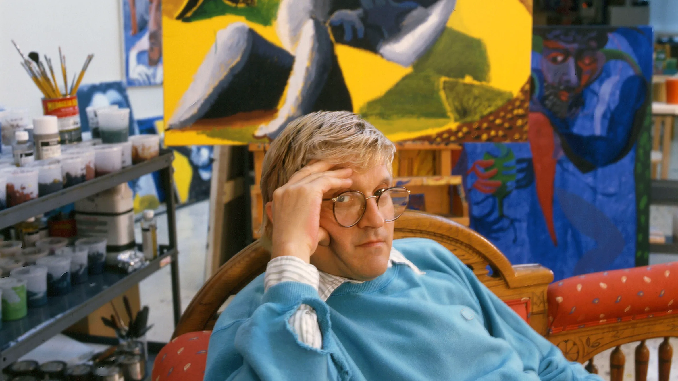 Hockney wearing a light blue sweatshirt and round glasses sits in a wooden chair inside a vibrant art studio. Behind them, large colorful paintings featuring expressive figures and bold yellow, blue, and green tones fill the space, while shelves of paint supplies and brushes line the left side of the room