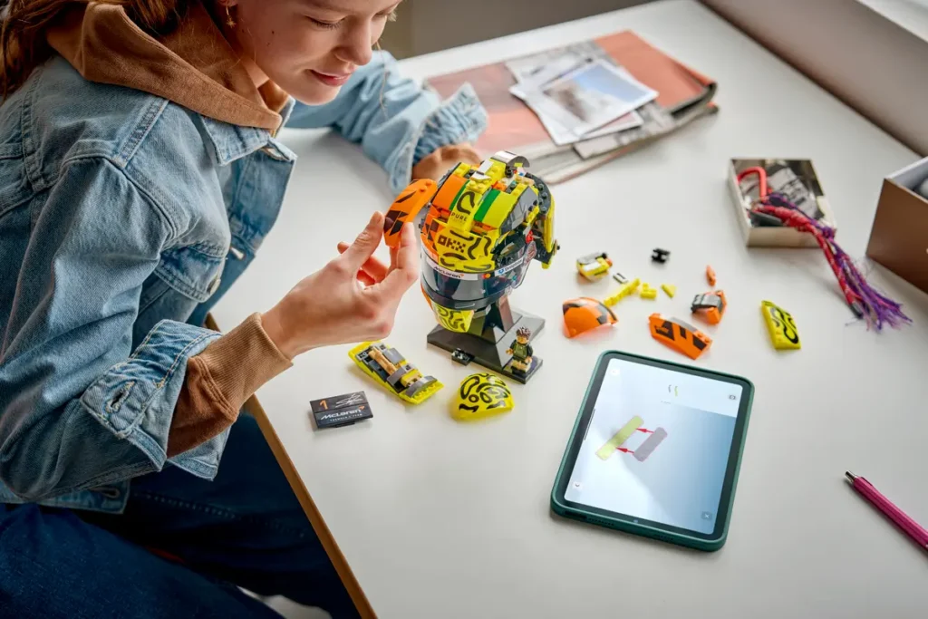 Builder constructing the LEGO Lando Norris helmet model using modular panels and a digital instruction guide on a tablet during the assembly process