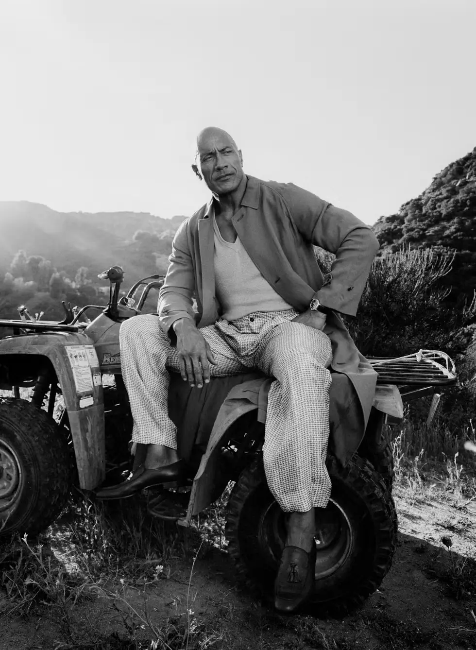 Dwayne Johnson in a striking black and white portrait, seated on an ATV in rugged terrain — wearing a tailored blazer and patterned trousers, embodying grounded strength and personal evolution in 2026