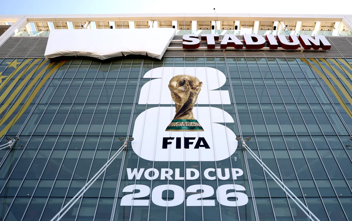 A large promotional banner for the 2026 FIFA World Cup covers the glass façade of a modern stadium. The display features the iconic FIFA World Cup trophy above bold “FIFA WORLD CUP 2026” branding, spanning multiple stories of the building. Above the banner, oversized red letters spell out “STADIUM,” while a large white-covered panel near the roofline suggests an upcoming reveal or installation. The image captures the scale and anticipation surrounding the international football tournament