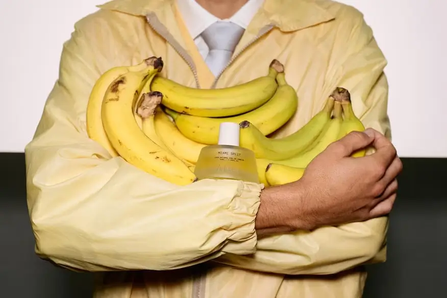 A person in a pale yellow jacket and white tie cradles a large bundle of ripe bananas, with a banana-themed fragrance bottle tucked among the fruit in a playful product-focused composition
