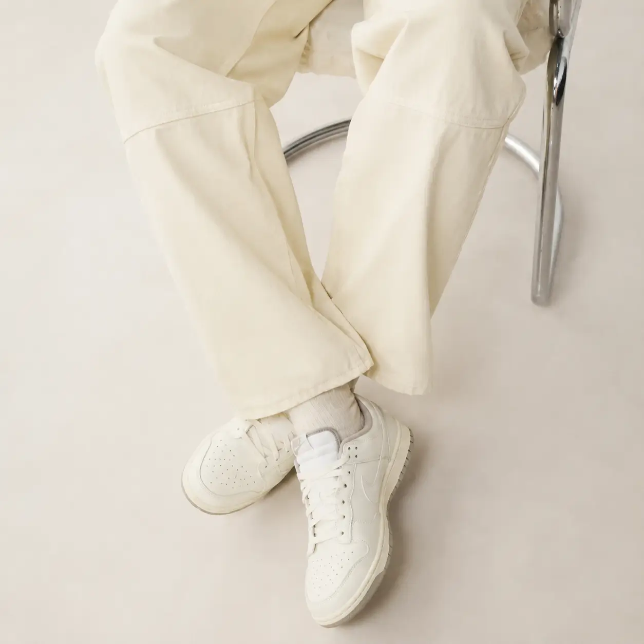 Person seated on a chrome chair wearing wide-leg cream trousers and matching socks, showcasing clean white Nike Dunk Low sneakers in a minimalist studio setting with a soft neutral background