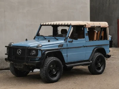 Light blue vintage Mercedes-Benz G-Wagen cabriolet parked in an industrial courtyard, featuring a beige soft top, black off-road wheels, rugged all-terrain tires, and military-inspired brush guards. The restored open-air 4x4 is photographed against a concrete wall, highlighting its classic boxy silhouette and handcrafted expedition-ready design