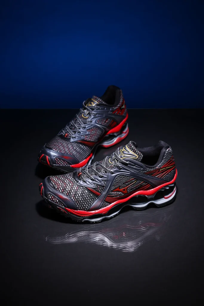 Studio product photograph of the Mizuno Wave Prophecy in a black and red colorway. The pair is displayed on a reflective black surface against a deep blue background, emphasizing the breathable mesh construction, metallic detailing, and distinctive Wave sole architecture