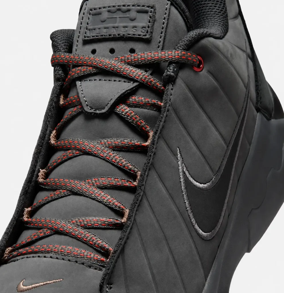 Close-up of the Nike x Matthew M. Williams low-top shoe in dark charcoal, highlighting the premium textured upper, oversized tonal Swoosh, red-speckled hiking-inspired laces, and subtle metallic mini Swoosh on the toe