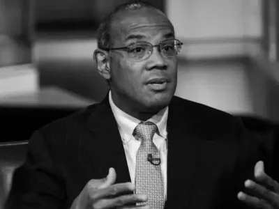 Black-and-white portrait of a man in a dark business suit and patterned tie speaking during an interview. Wearing glasses and gesturing with both hands, he appears engaged in conversation while seated in a studio setting with a softly blurred background