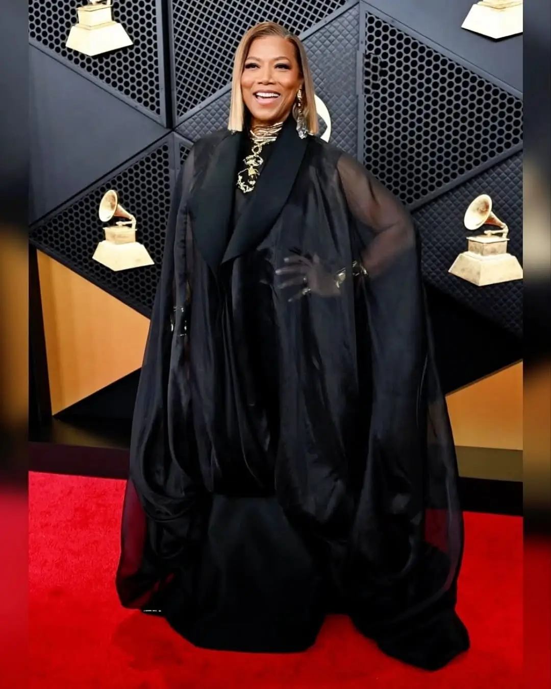 A celebrity attends a red carpet event wearing a dramatic oversized black gown with flowing sculptural sleeves, sheer accents, and layered gold jewelry, posing in front of a backdrop featuring gramophone trophy motifs and geometric panel