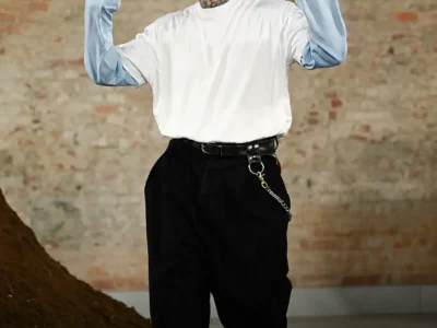 A fashion runway model walks confidently through an industrial-inspired show space with exposed brick walls and a dark textured floor. The model wears an oversized white graphic top layered over pale blue sleeves, wide black trousers with a chain accessory, black footwear, sunglasses, and a black baseball cap, while neck tattoos and raised hands add a play, expressive touch