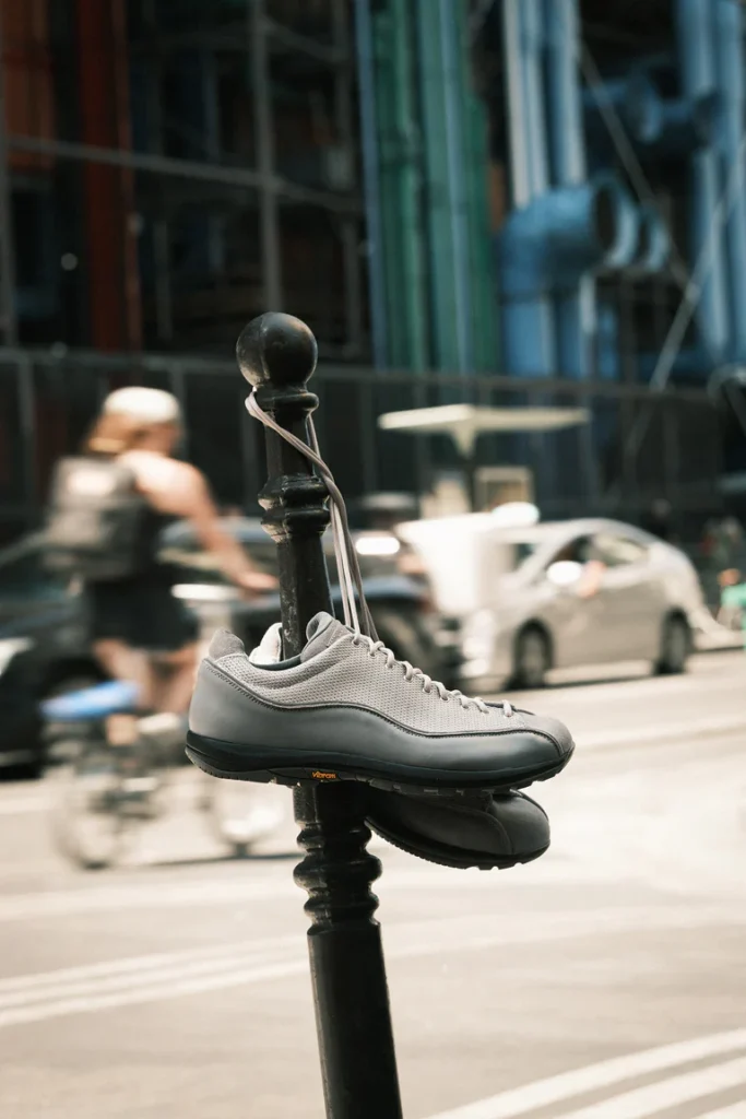 Gray DIEMME low-top sneakers suspended from a black street bollard by their laces, with cyclists, cars, and a modern city streetscape softly blurred in the background